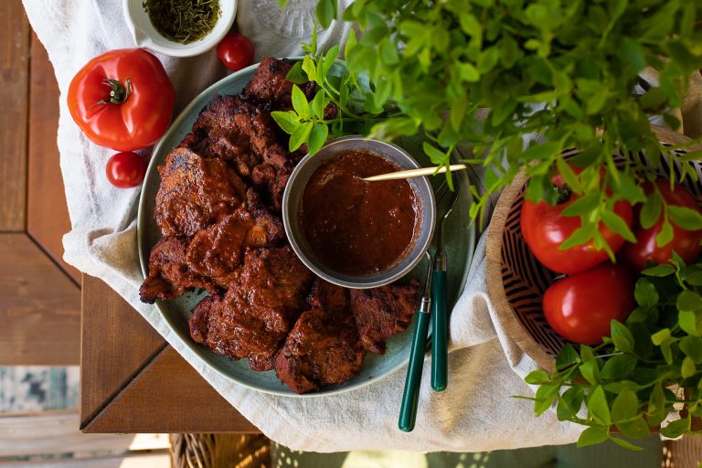 Pruunil laual naturaalset linast beežikat tooni laualina, millele on asetatud tomatid ja väike kauss kuivatatud ürtidega. Foto keskel fookuses on suur ümmargune rohekas taldrik, millel on näha punase marinaadiga lihatükid. Taldrikule lihatükkide kõrvale on asetatud kauss marinaadiga, millesse on kuldne lusikas sisse torgatud. Taldrikule toetuvad ka rohelise sabaga söögiriistad. Pildi paremas üleval servas on roheline taim ning all servas musta-valgekirju puitkauss suurte tervete tomatitega.