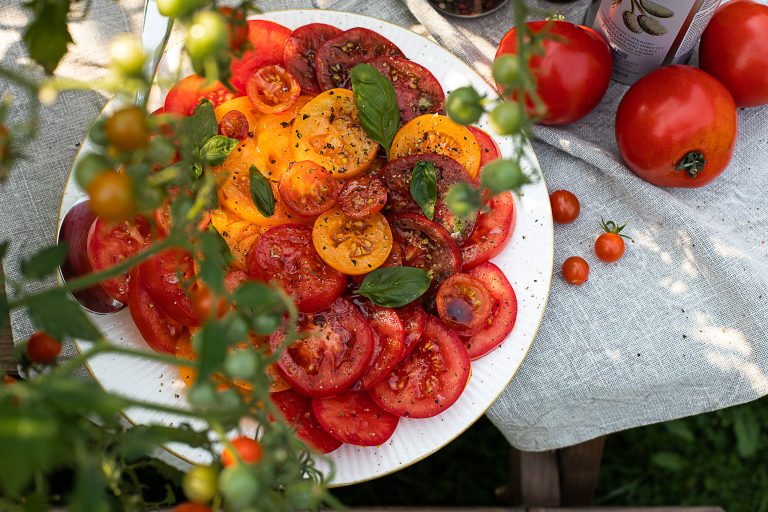 Suvine õues tehtud toidufoto, kus murule asetatud puitlaud on kaetud linase heleda laudlinaga. Sellele on asetatud suuri ja väikeseid terveid tomateid. Vasakul servas on uduselt tomatitaim. Fookuses on laual suur valge taldrik, millele on asetatud virn punaste ja kollaste tomatite viile. Sellele on peale valatud tumedat kastet ja raputatud maitseaineid. Salat on kaunistatud basiilikulehtedega.