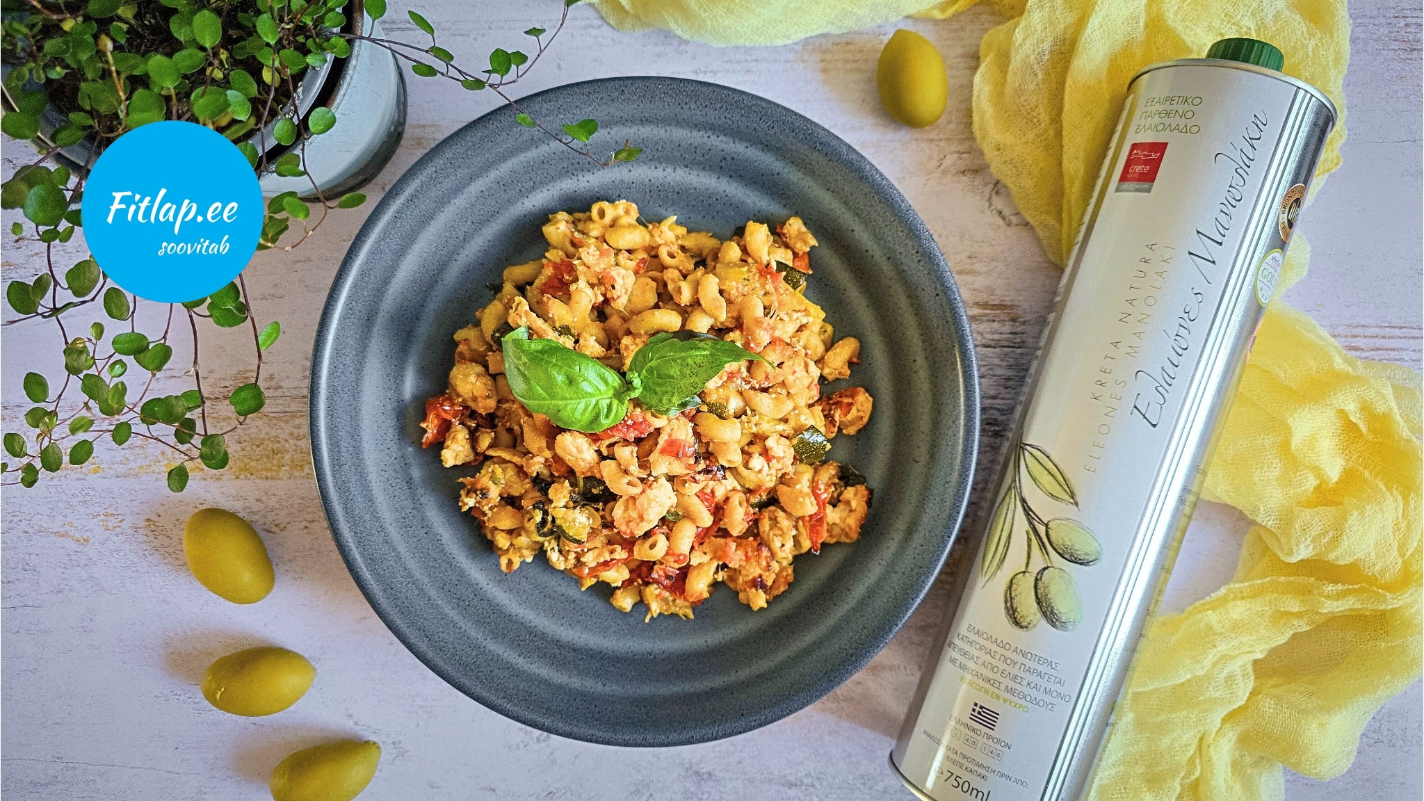 Puidust laual on hallikassinine taldrik, millel on mis sisaldab tomatit ja suvikõrvitsat. Kaunistuseks on sellele pandud värske basiilikuoks. Pildi vasakus ülemises servas on roheline taim, mille all on kolm rohelist oliivi. Taldrikust paremal on lauale diagonaali asetatud valges plekkupudelis oliiviõli, mille alla on laotatud kollane volditud kangas. Pildil vasakus servas on sinisel taustal logo, mille sisse on kirjutatud fitlap.ee soovitab.